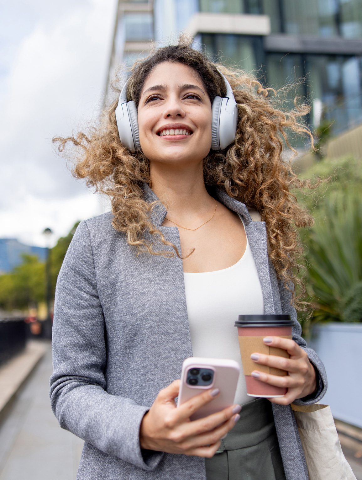 Cheerful young woman with long, curly hair wearing white over-ear headphones, a gray blazer, and a white top. She is standing outdoors, holding a coffee cup in one hand and a pink smartphone in the other, smiling broadly with an urban background of buildings and greenery.
